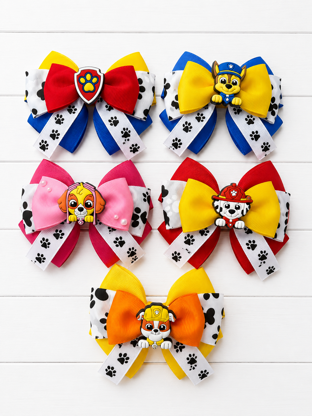 Six colorful Paw Patrol hair bows arranged on a white wood background. Each bow features a large color-blocked top section with red, yellow, blue, pink, or orange, a Paw Patrol character charm in the center, and white ribbon tails printed with black paw prints.