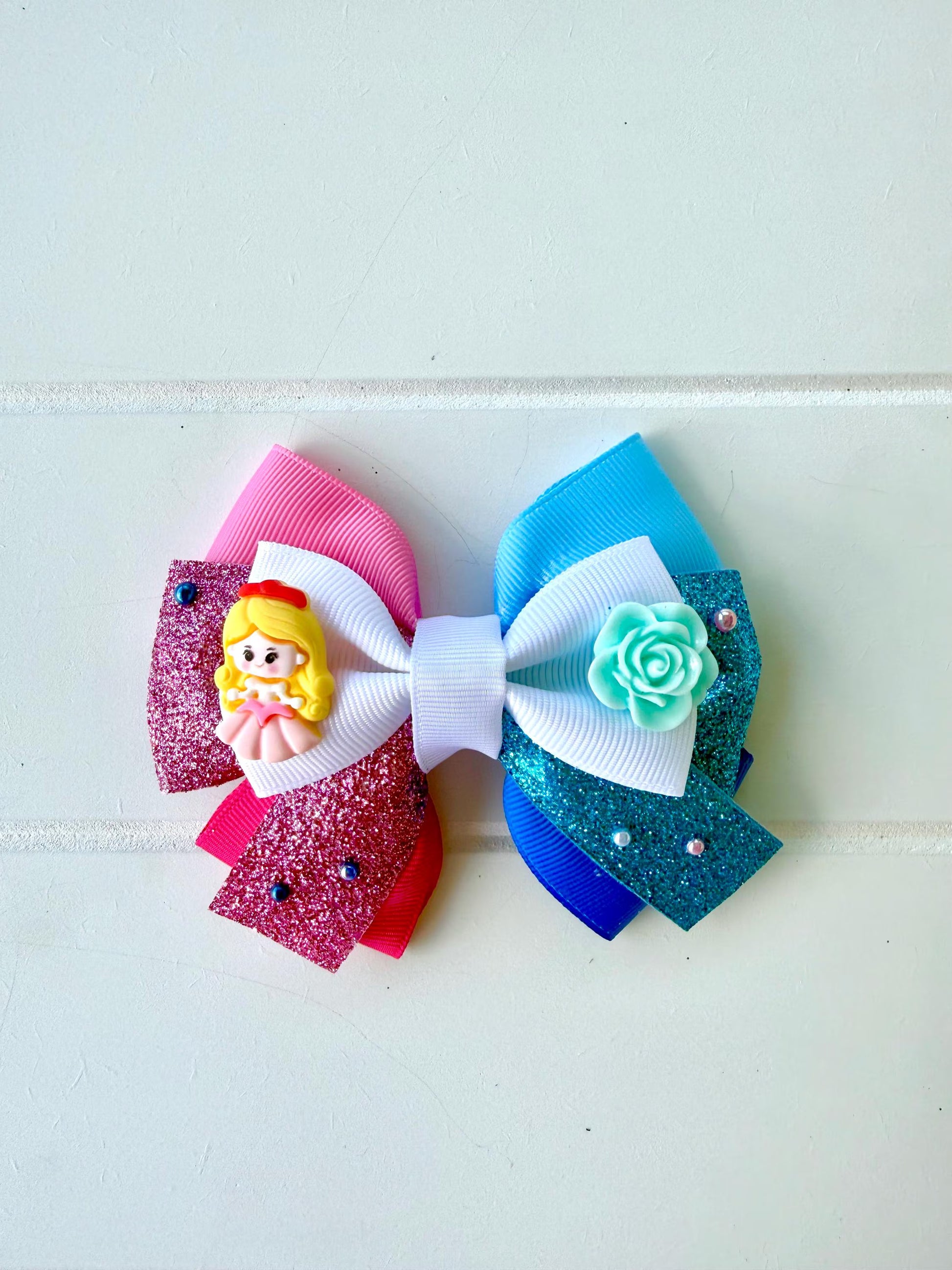 A handmade hair bow featuring multiple layers of pink, blue, and white ribbons, embellished with a small yellow princess charm and a mint green rose, set against a light off-white paneled background.