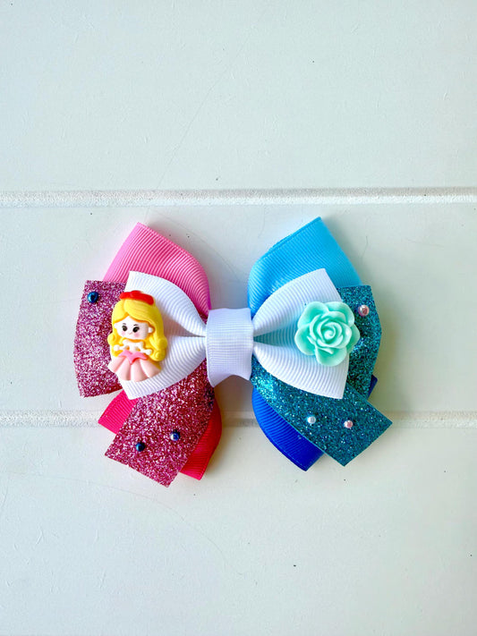 A handmade hair bow featuring multiple layers of pink, blue, and white ribbons, embellished with a small yellow princess charm and a mint green rose, set against a light off-white paneled background.