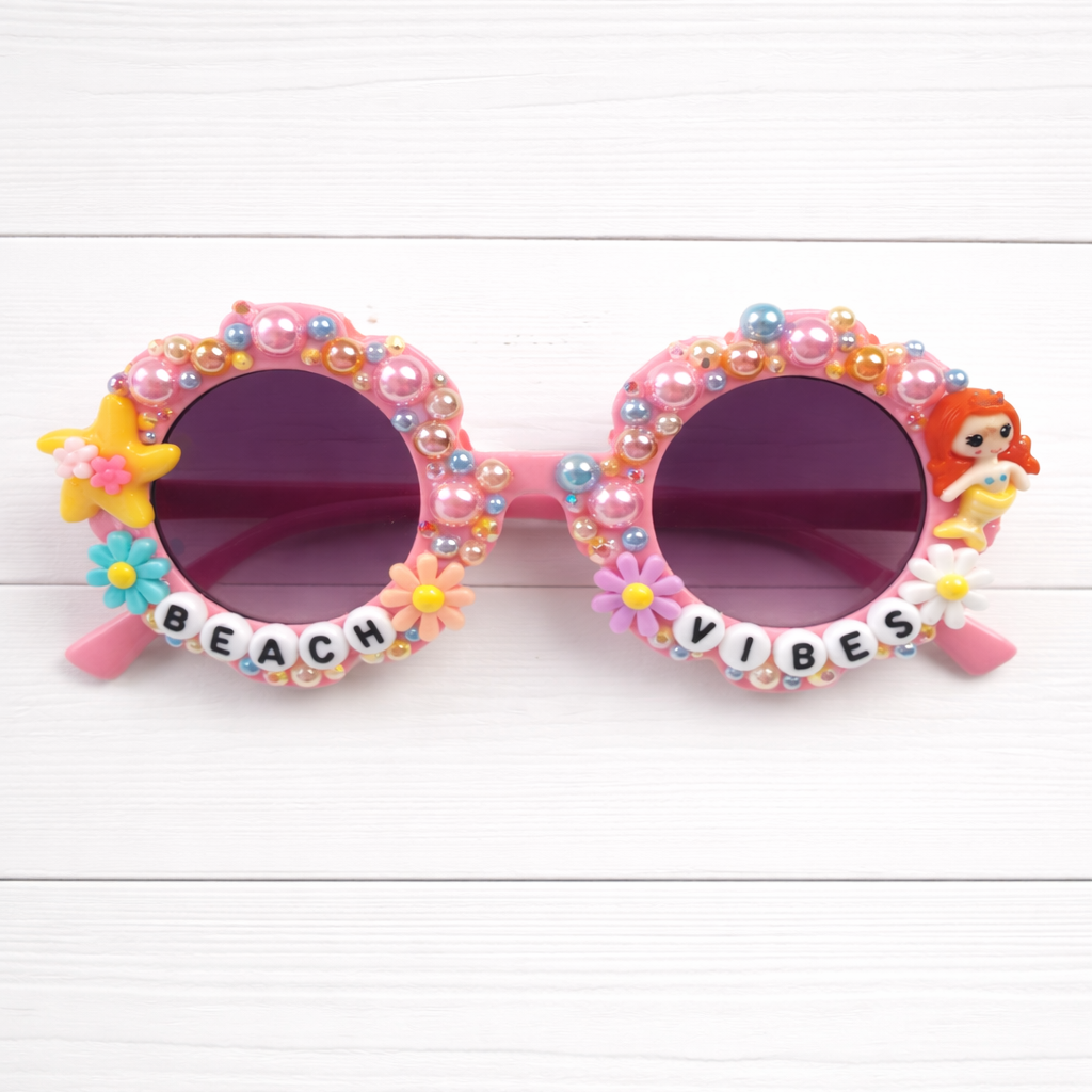 Kids flower pink Sunglasses with colorful bead embellishments and 'Beach Vibes' text on a white wooden background. Mermaid charm on right and starfish charm on left.