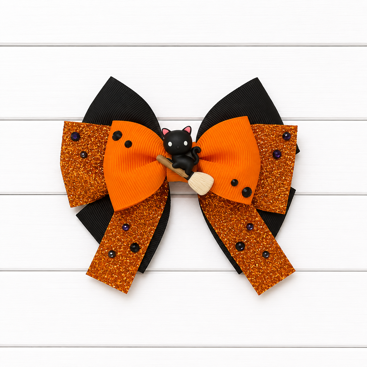 A double-layered hair bow featuring orange grosgrain ribbon, orange glitter panels with black bead accents, black ribbon loops, and a small black cat wearing a witch hat on a broomstick at the center, displayed on a white wood plank background.