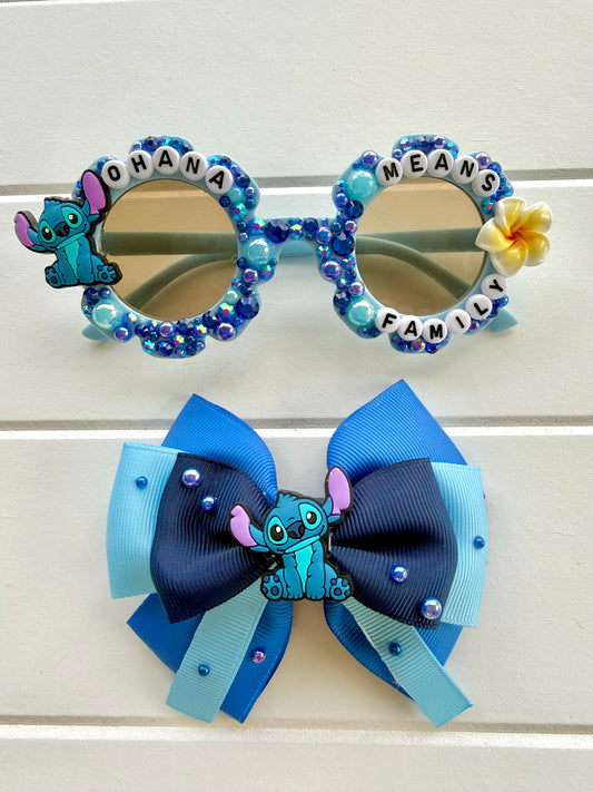 Custom blue and light blue rhinestone-studded novelty sunglasses with a blue flower accent and a matching blue ribbon hair bow featuring a Stitch character, displayed on a white paneled background.