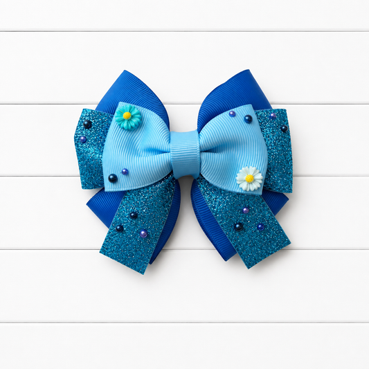 A blue hair bow featuring light blue grosgrain ribbon, teal glitter panels, small white daisy embellishments, and blue beads, displayed on a white wood plank background.