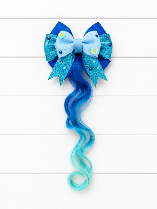 A decorative hair bow accessory with layered blue and turquoise glitter and fabric bows, small daisy embellishments, and a long wavy hair extension in blue fading to aqua, displayed on a white wood plank background.