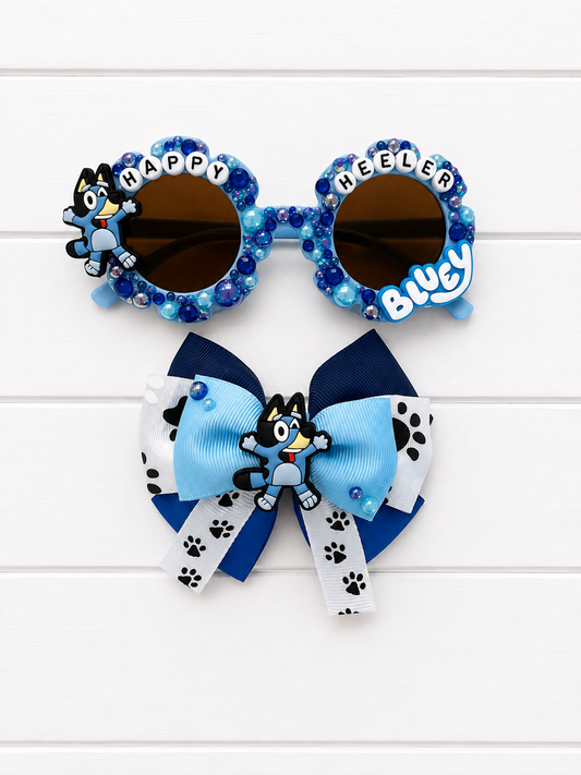 A pair of novelty sunglasses and a matching hair bow set, both themed around Bluey. The sunglasses feature blue rhinestone frames and a Bluey charm on the left with 'HAPPY HEELER' letters. The bow features a Bluey charm in the middle and navy, blue, light blue and paw print ribbon. 