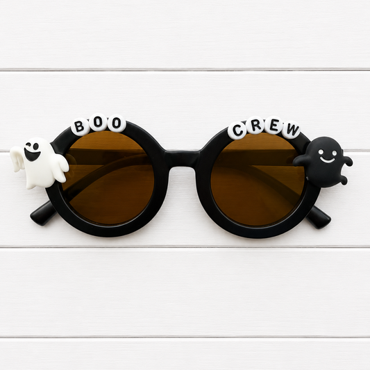 Black round sunglasses with brown lenses, decorated with a white ghost on the left, a black ghost on the right, and the words 'BOO CREW' across the top of the frames, displayed on a white wood plank background.