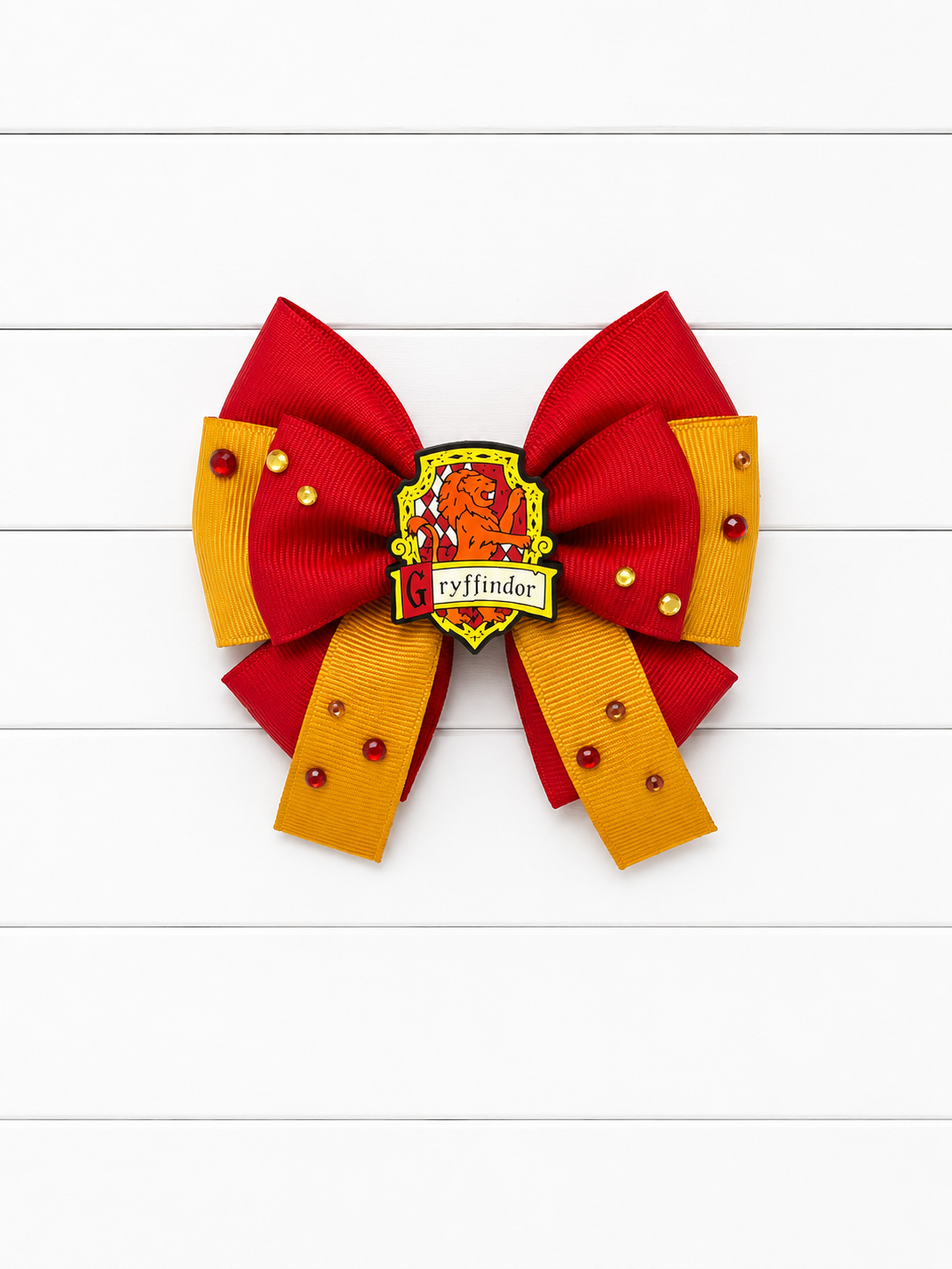 A red and gold ribbon hair bow featuring a Gryffindor crest pin in the center and small red rhinestone accents, displayed on a white wood plank background.