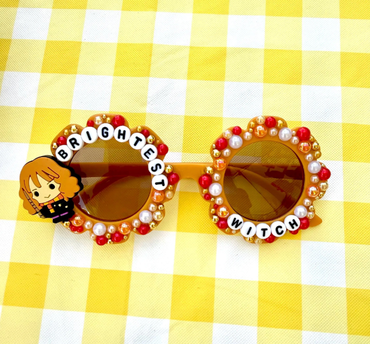 Kids brown flower sunglasses with Hermione charm on left and "Brightest Witch" text around the frames. There are red gold and white pearls outlining the whole frames.