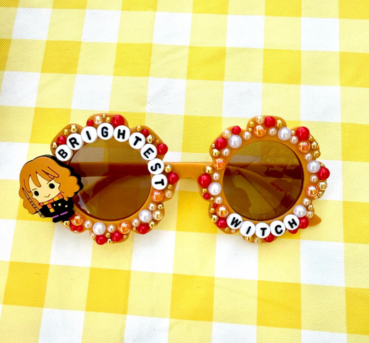Kids brown flower sunglasses with Hermione charm on left and "Brightest Witch" text around the frames. There are red gold and white pearls outlining the whole frames.