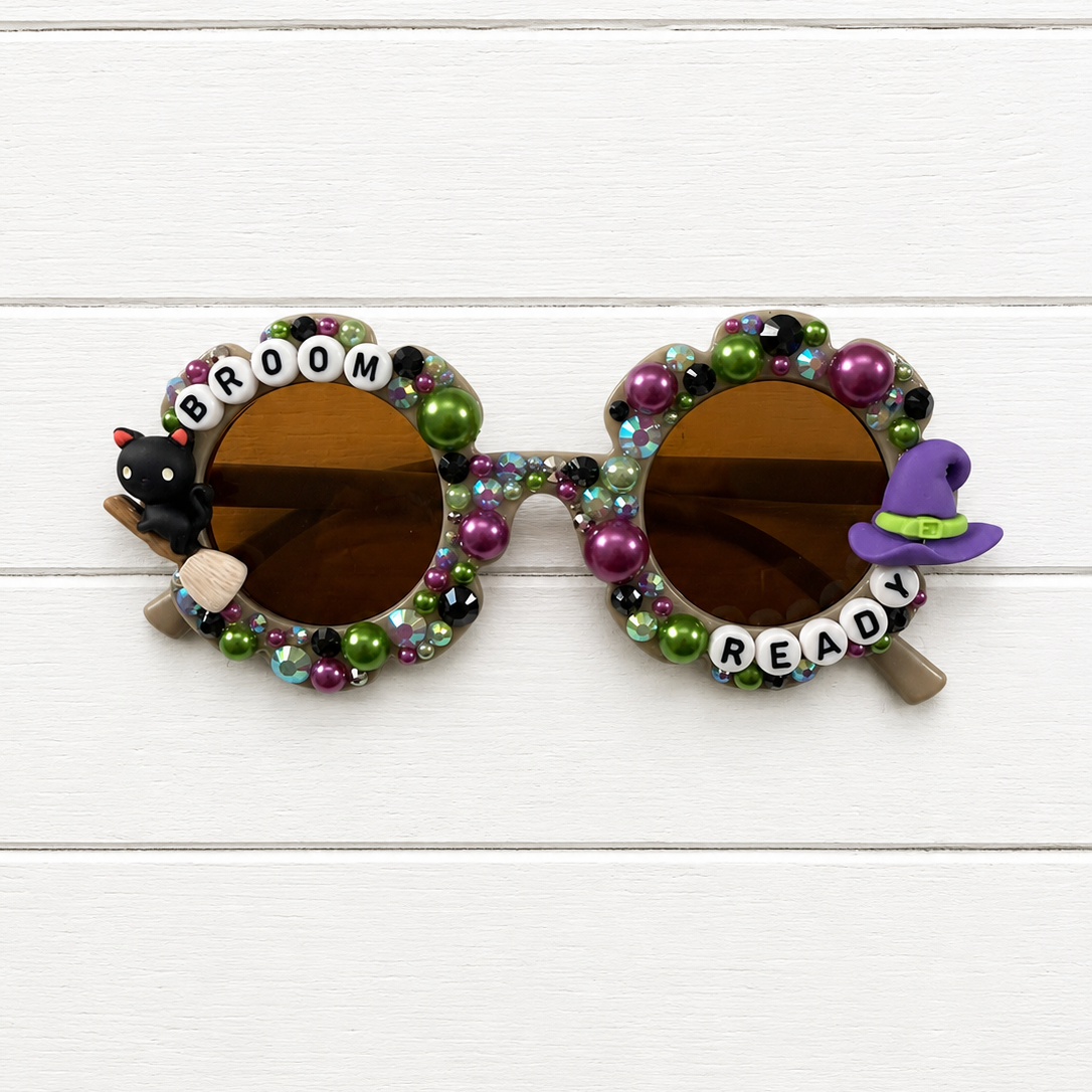 Decorated round sunglasses with brown lenses, covered in colorful beads and small charms including a black cat, a purple witch hat, and letter beads spelling 'BROOM READY' on a white wood plank background.