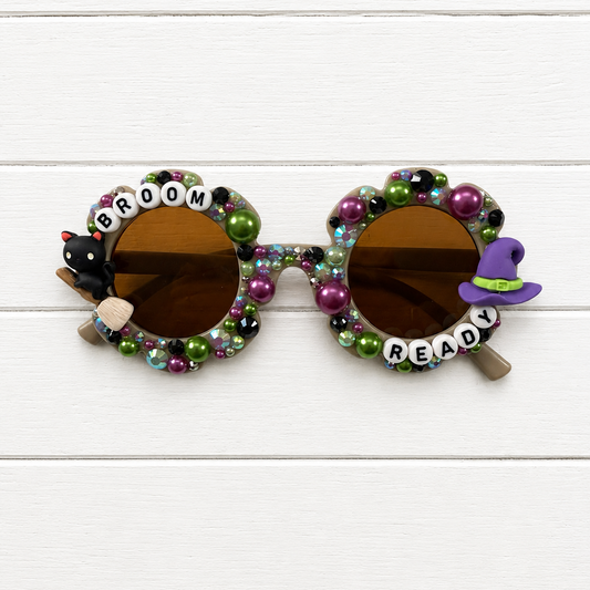 Decorated round sunglasses with brown lenses, covered in colorful beads and small charms including a black cat, a purple witch hat, and letter beads spelling 'BROOM READY' on a white wood plank background.