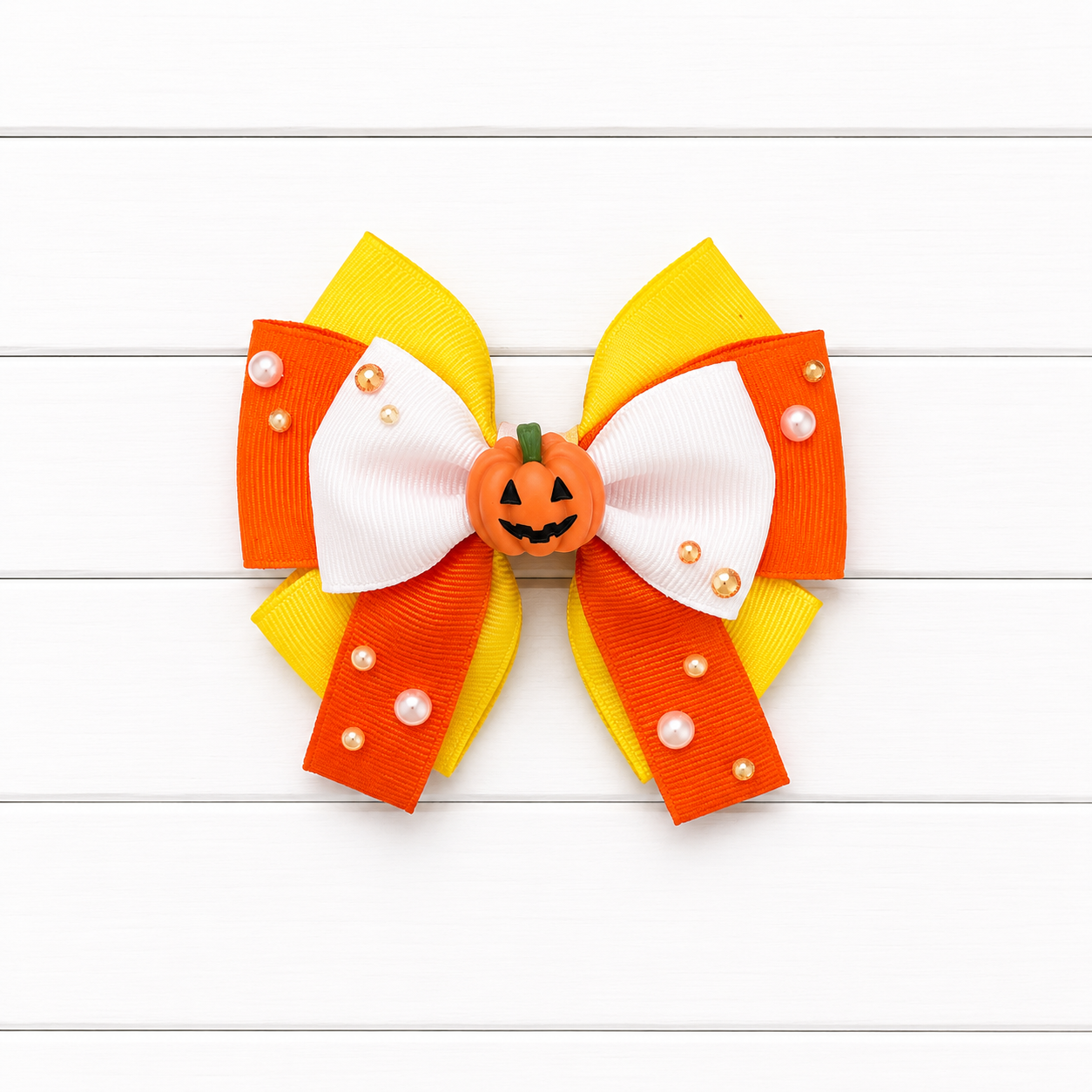 A Halloween-themed hair bow accessory featuring orange, yellow, and white ribbons styled like candy corn, with a small jack-o'-lantern ornament in the center and decorative pearl-like beads, shown on a white wood plank background.
