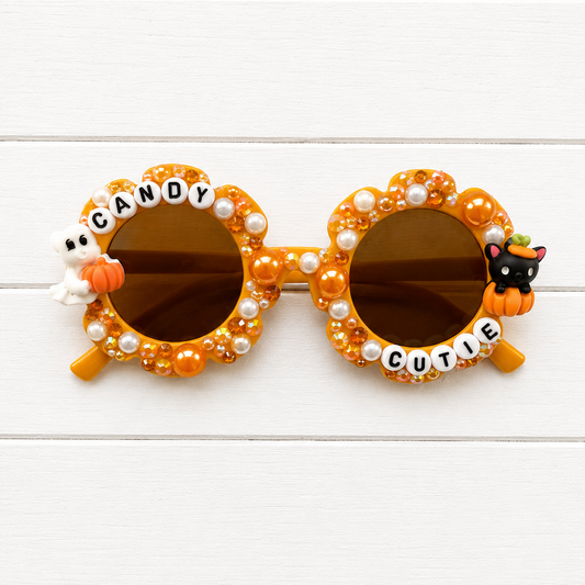 Candy Corn themed novelty sunglasses with orange frames decorated with faux pearls, orange beads, letter tiles spelling 'CANDY' and 'CUTIE', a white ghost on the left, and a black cat on the right, shown on a white wood background.