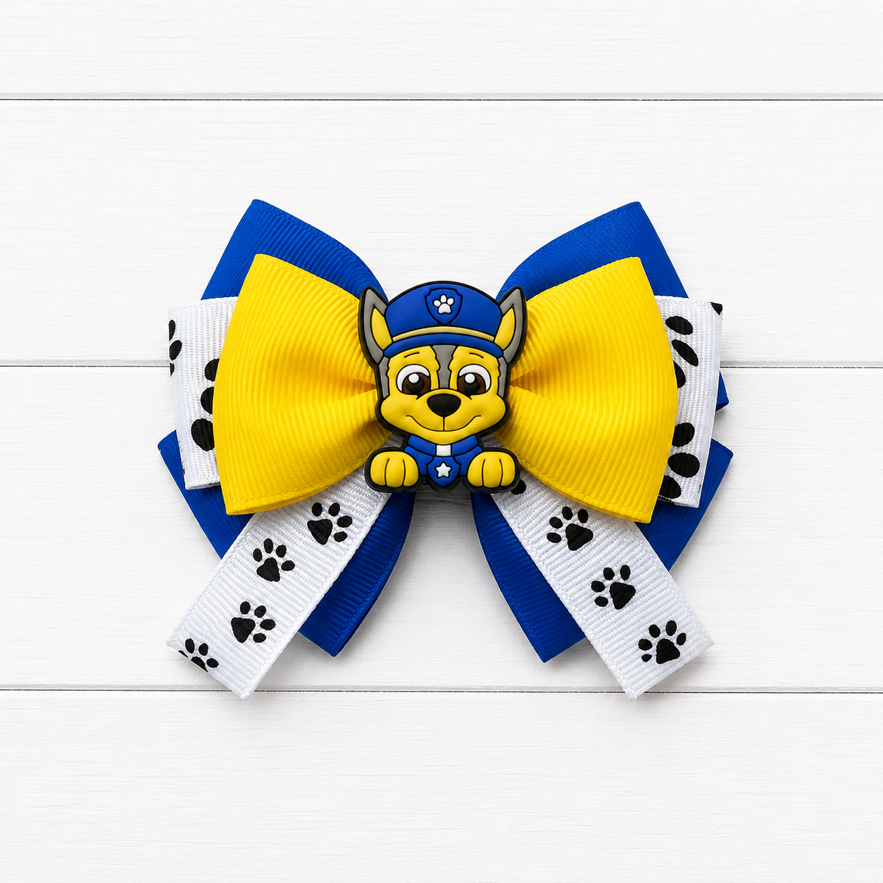 A girl's hair bow featuring a yellow and blue grosgrain ribbon design with Paw Patrol character Chase in the center, set against a white wood plank background.