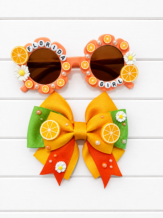 A pair of novelty sunglasses that say "FLORIDA GIRL" with oranges around the frames and a matching hair bow set with orange, yellow, and green citrus-themed decorations, displayed on a white wood background.