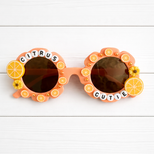 Kids flower orange sunglasses with two orange charms and "Citrus Cutie" text surrounding the frames. Little orange embellishments and orange pearls outline the frames.