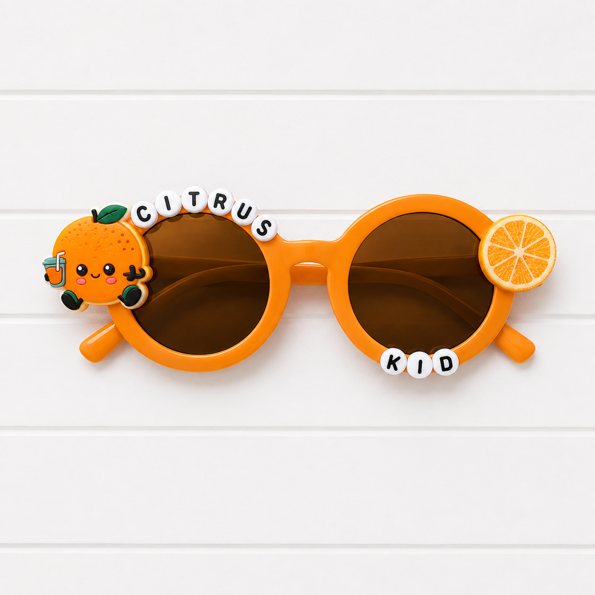 Bright orange round children's sunglasses with dark brown lenses, decorated with a cute cartoon orange character on the left frame, a small orange slice charm on the right, and white letter tiles spelling 'CITRUS KID' above and below the frame, shown on a white wood background.