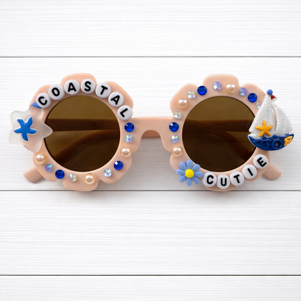 Kids light pink flower sunglasses with "Coastal Cutie" text surrounding the lens and a sailboat charm on the right. There are blue and pink rhinestones outlining the frames.