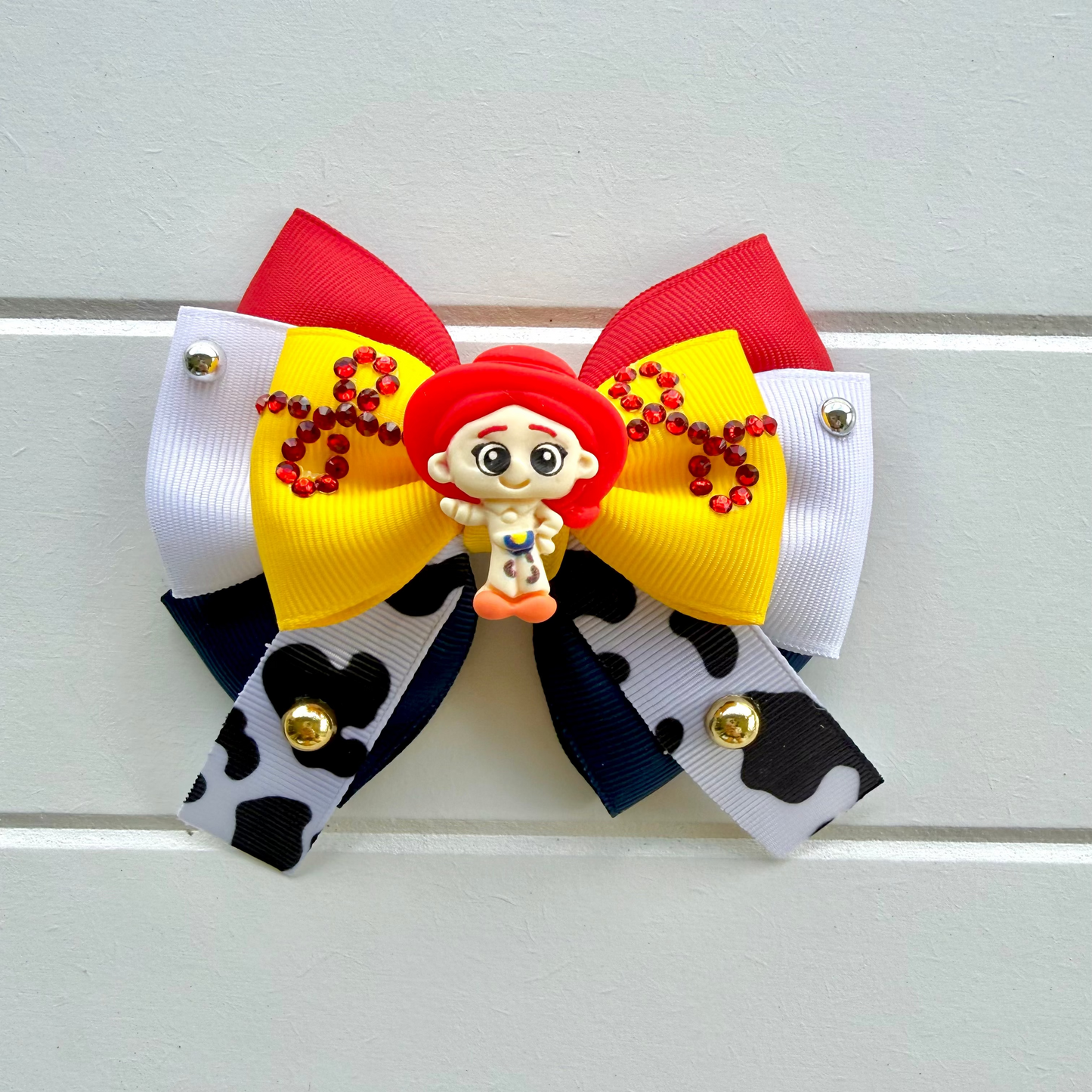 A decorative hair bow featuring a red and yellow ribbon top with red embroidered lettering, a small Jessie haracter charm in the center, and a bottom layer of white and black cow-print ribbon with small gold bells, shown against a light gray paneled background.