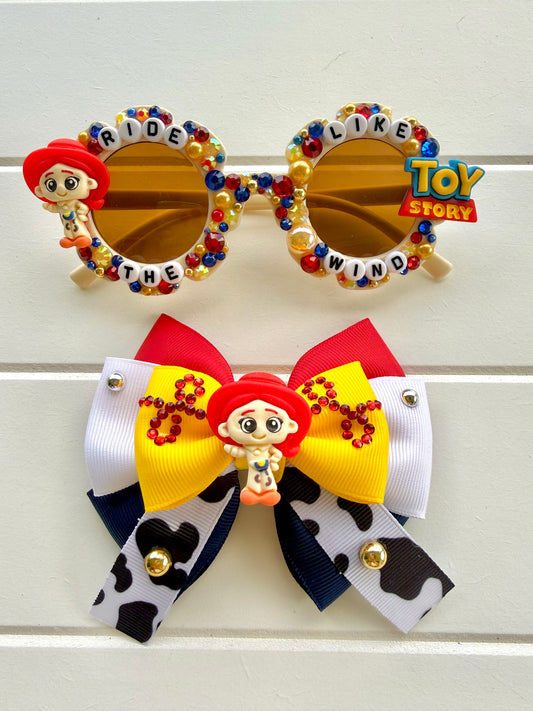 Matching sunglasses and bow. Sunglasses feature a "Jessie" charm on the left and "Toy Story" charm on the right. Yellow, blue, and red rhinestones and pearls and text "RIDE LIKE THE WIND" outlines the frames. The bow is red, white, navy, yellow, and cow ribbon with a Jessie charm in the middle.
