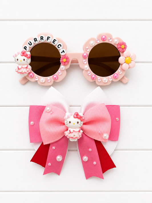 Pink Hello Kitty themed hair accessories set, including decorative round sunglasses with flower and pearl embellishments and a large pink and red ribbon bow with a Hello Kitty charm.