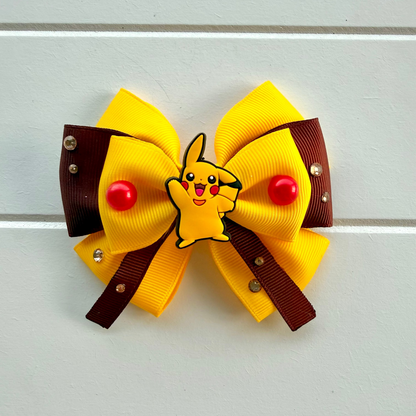 A yellow and brown hair bow featuring a small Pikachu character in the center, with red pom-poms and rhinestone accents, displayed on a white paneled background.