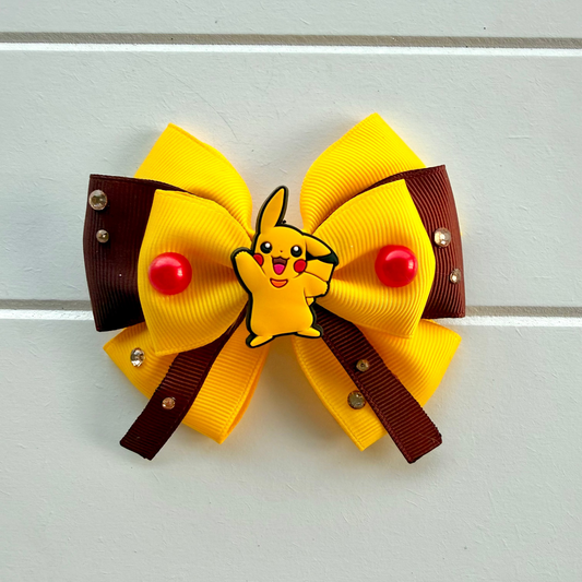 A yellow and brown hair bow featuring a small Pikachu character in the center, with red pom-poms and rhinestone accents, displayed on a white paneled background.