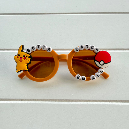 Kids brown round sunglasses with decorative bead lettering and character accents, featuring Pikachu on one lens and a red and white Pokéball on the other, displayed on a white paneled background.