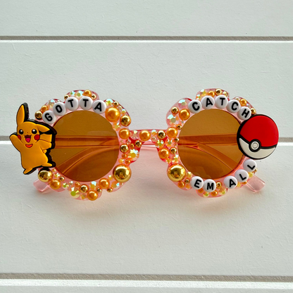 Decorative round sunglasses with amber-tinted lenses, pink frames, rhinestone embellishments, a Pikachu character on the left lens rim, and a Pokéball on the right lens rim, displayed on a white wood background.