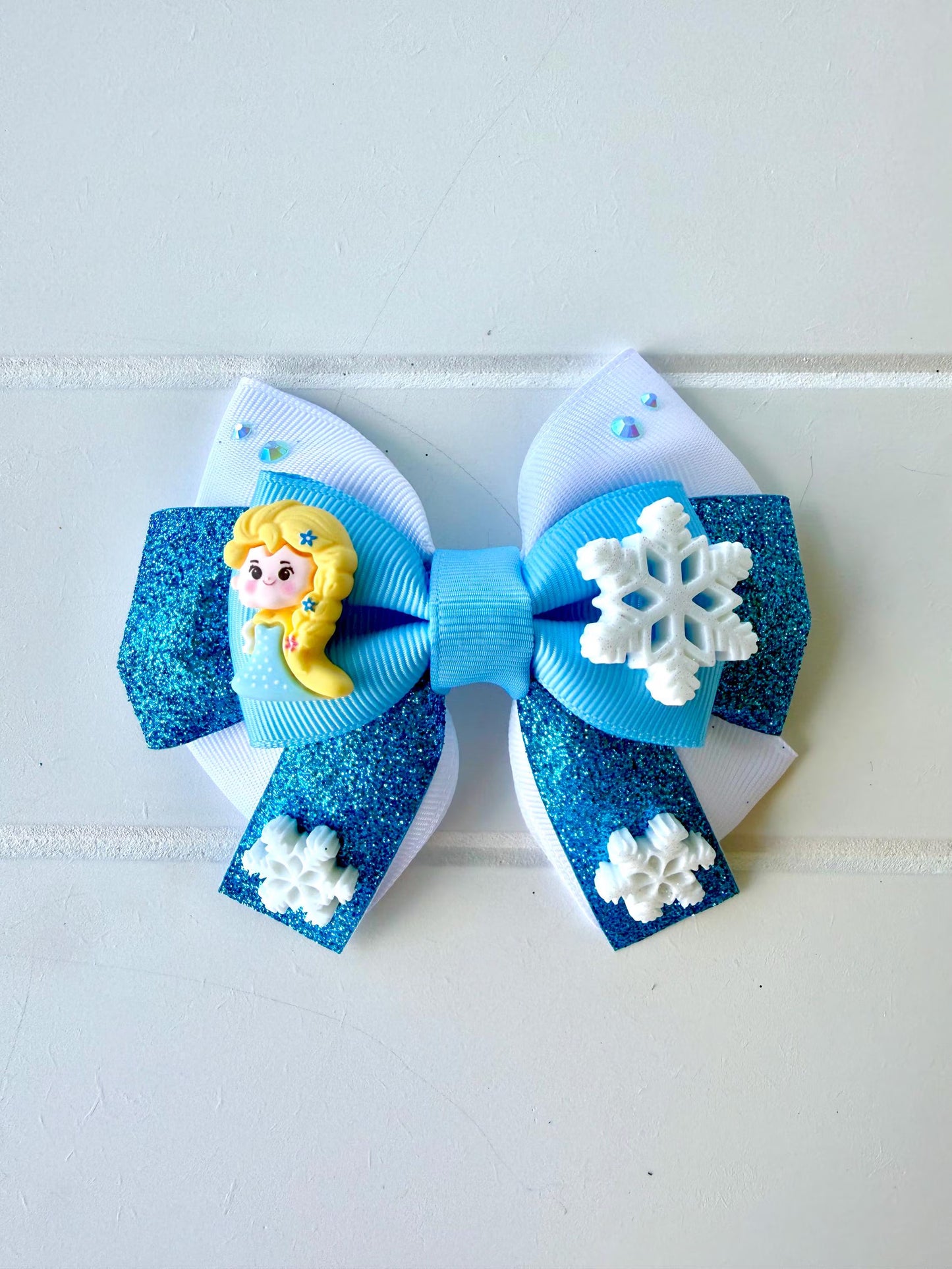 A hair bow featuring light blue and white ribbons, decorated with a blue glitter section, white snowflakes, and a yellow-haired Elsa charm, displayed on a white paneled background.