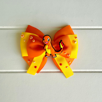A hair bow featuring a double-loop design in orange and yellow grosgrain ribbon, embellished with small clear rhinestones and a central Charmander character charm, displayed against a white paneled background.