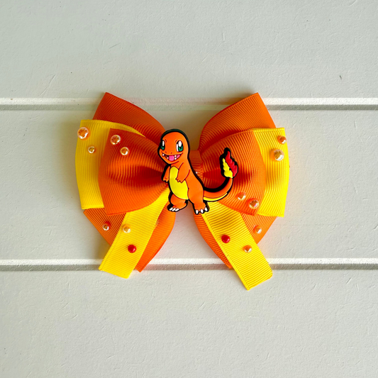 A hair bow featuring a double-loop design in orange and yellow grosgrain ribbon, embellished with small clear rhinestones and a central Charmander character charm, displayed against a white paneled background.