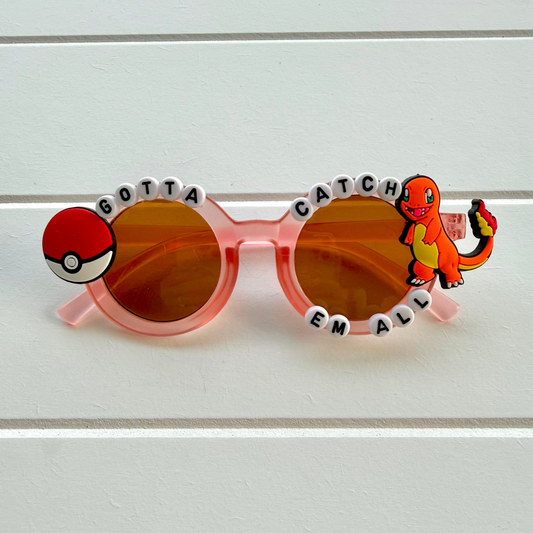 Red transparent kids round sunglasses with orange lenses, featuring a Pokéball charm on the left and a Charmander charm on the right, with letter beads spelling 'GOTTA CATCH EM ALL' around the frame.
