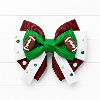 A decorative hair bow featuring a green center and loops, brown side panels, white ribbon tails, small green and pearl rhinestones, and two small football embellishments on the front, shown on a white wood background.