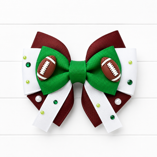 A decorative hair bow featuring a green center and loops, brown side panels, white ribbon tails, small green and pearl rhinestones, and two small football embellishments on the front, shown on a white wood background.