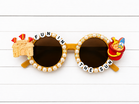 Kids brown round sunglasses with sandcastle charm on left and bucket of sand charm on right. "FUN IN THE SUN" letters outline the frames and yellow pearls and rhinestones decorate the frames.