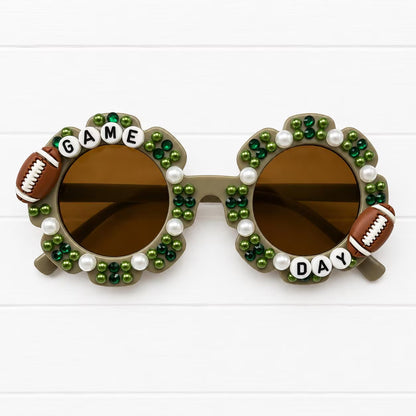 Kids grey flower sunglasses with two football charms and green and white pearls and rhinestones decorating the frames. "GAME DAY" text outlines the frames.