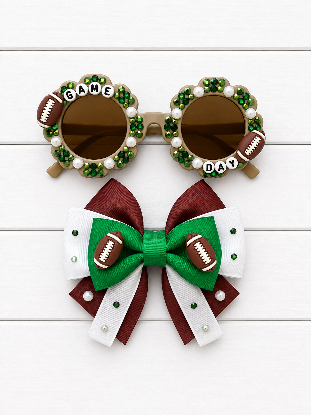 A pair of novelty football-themed sunglasses and a matching hair bow set on a white wood background. The round sunglasses have brown lenses, green and white rhinestone borders, faux football details, and the words 'GAME DAY' on the bridge.