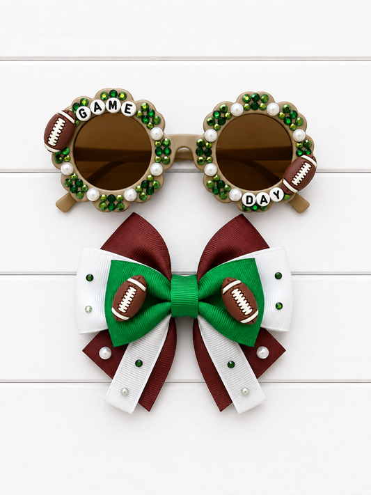 A pair of novelty football-themed sunglasses and a matching hair bow set on a white wood background. The round sunglasses have brown lenses, green and white rhinestone borders, faux football details, and the words 'GAME DAY' on the bridge.