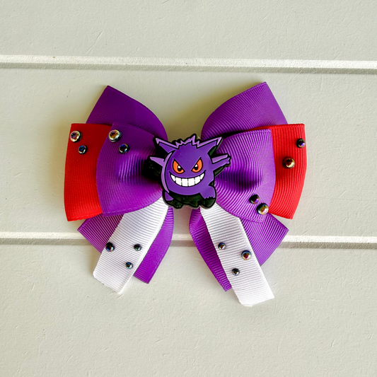 A hair bow featuring a purple and red ribbon design with white ribbon tails, small black rhinestones, and a Gengar charm, displayed on a light beige paneled background.