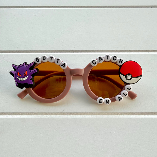 Round purple sunglasses with amber-tinted lenses, decorated with a Gengar charm on the left and a red-and-white Pokéball charm on the right, with white letter beads spelling 'GOTTA CATCH EM ALL' across the top and bottom rims, shown on a white paneled background.