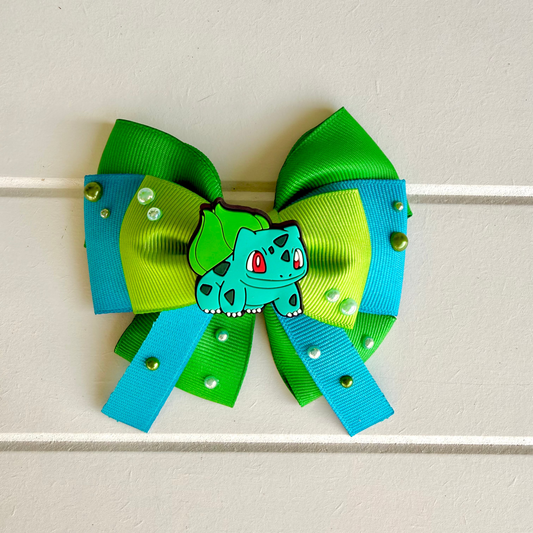 A handmade hair bow featuring bright green and turquoise ribbons, adorned with small rhinestones and metallic studs, and decorated with a central green and blue Bulbasaur character charm.