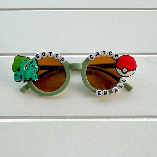 Light green novelty sunglasses with brown lenses, decorated with a Bulbasaur charm on the left and a Pokéball charm on the right, separated by white letter beads spelling 'GOTTA CATCH EM ALL'.