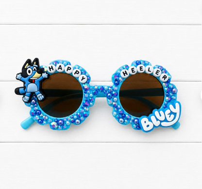 Children's sunglasses with blue frame and Bluey charm on left and "HAPPY HEELER" letters and blue pearls and rhinestones outlining the frames.