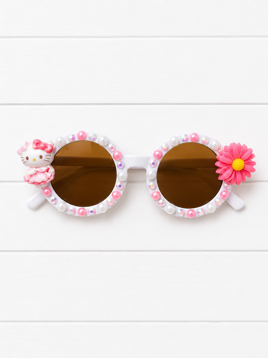 Decorative round sunglasses with brown lenses, white frames adorned with pink and white pearls, a Hello Kitty figure on one side, and a pink flower on the other, displayed on a white wood background.