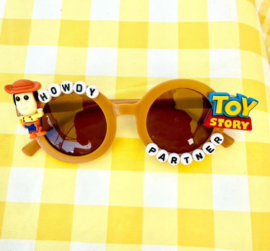 Kids round brown sunglasses with Woody charm on left and Toy Story charm on right. "Howdy Partner" text oulines the frames.