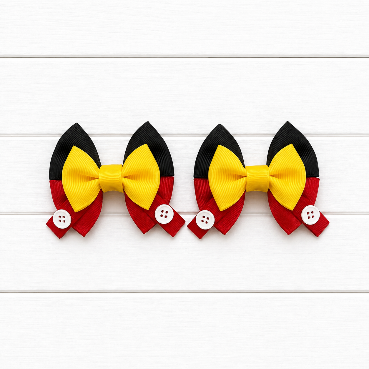 Two matching hair bows arranged side by side on a white wood plank background. Each bow features a large yellow center with black upper wings and red lower wings, plus a small white button detail on the red side.