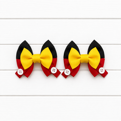 Two matching hair bows arranged side by side on a white wood plank background. Each bow features a large yellow center with black upper wings and red lower wings, plus a small white button detail on the red side.
