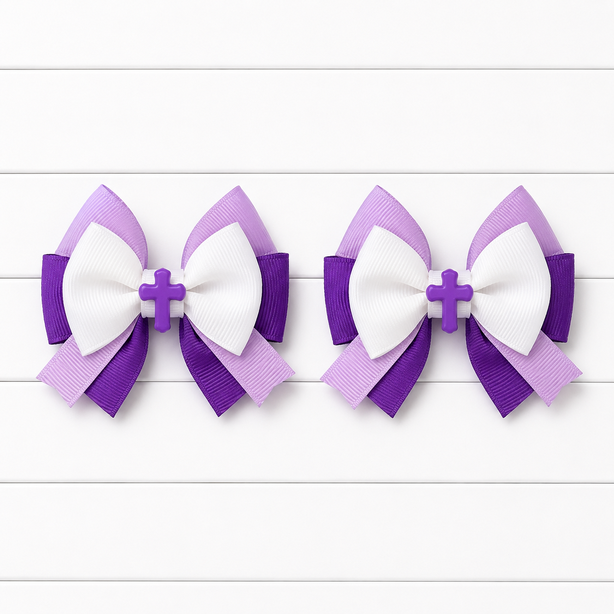 Two matching purple and white ribbon hair bows with a small cross center, displayed side by side on a white wood plank background.