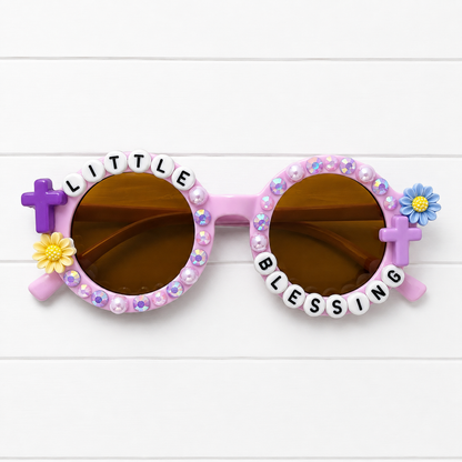 Light purple kids round sunglasses with brown lenses, decorated with rhinestones, white letter beads spelling 'LITTLE BLESSING', purple crosses, and small yellow and blue flowers on a white wood background.